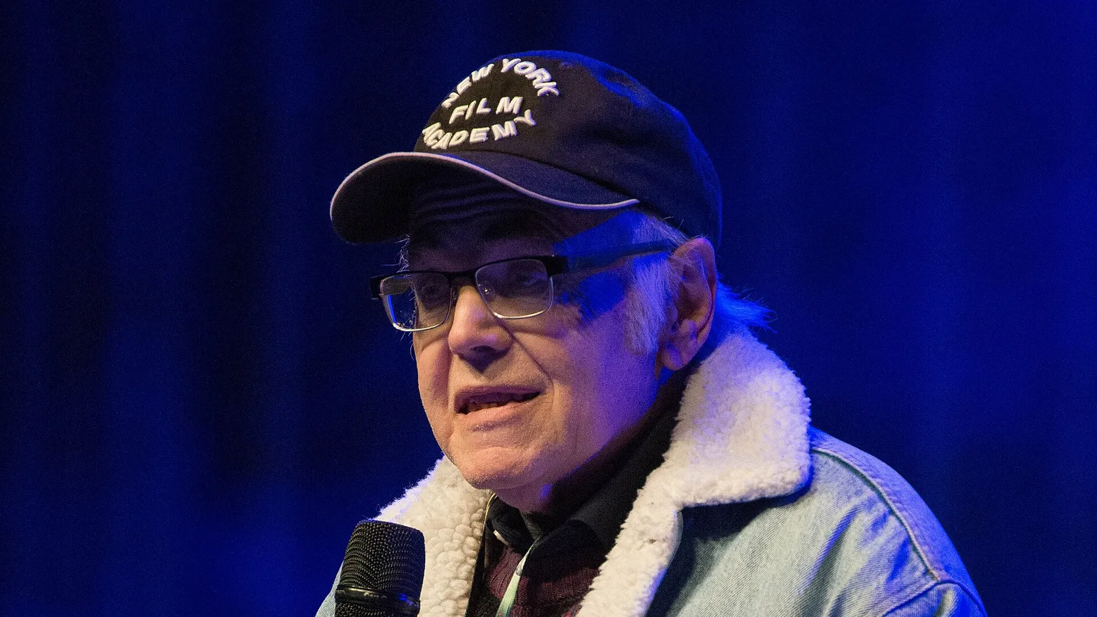 Walter Koenig speaking into a microphone on stage at a fan convention in 2016, wearing a cap and a Star Trek patch on his denim jacket